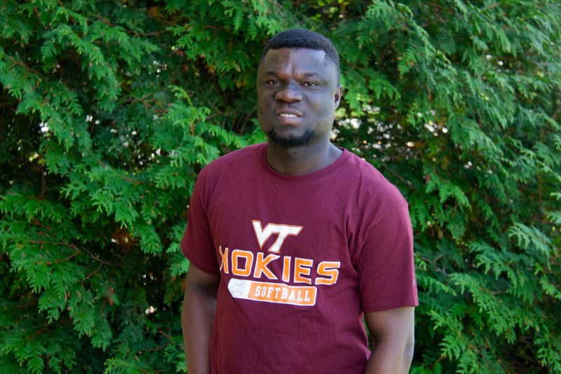 A man wearing a Virginia Tech t-shirt stands in front of a hedge.