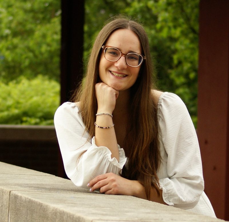 A woman with long dark hair and glasses smiles as she leans against a concrete wall.