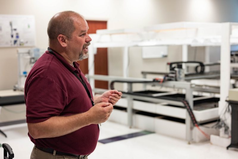 Researcher standing with tables that incorporate robotics, technology, and camera images to determine plant health.