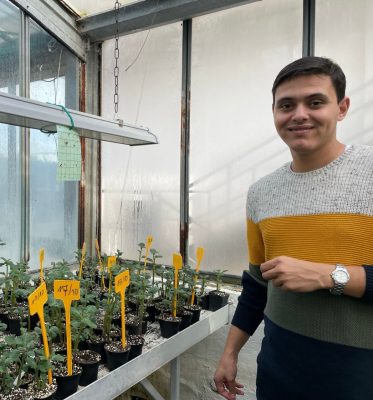 A young man stands beside a table full of potted plants with yellow markers in a greenhouse.