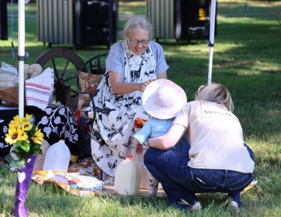 A woman in a black and white cow apron smiles at a young child and her mother