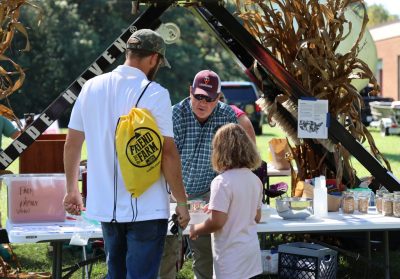 A man in a plaid shirt speaks to another man and a young child at a table with corn stalks