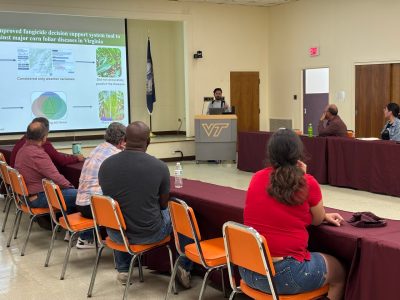 A young man delivers a presentation from behind a Virginia Tech podium while a large screen is displayed beside him.
