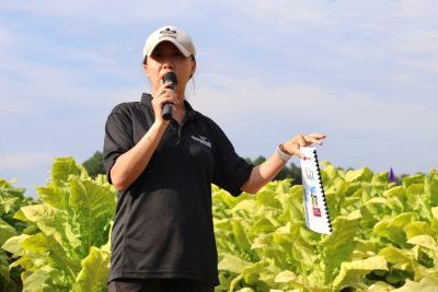A woman in a black shirt speaks into a microphone beside a tobacco field.