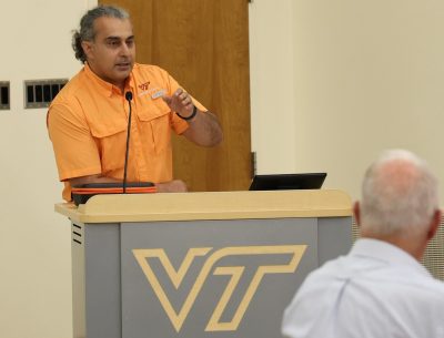 A man in an orange shirt speaks into a microphone from behind a podium