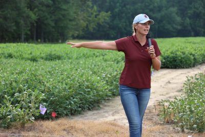 A woman in a maroon shirt speaks into a microphone as she points to a field of soybeans