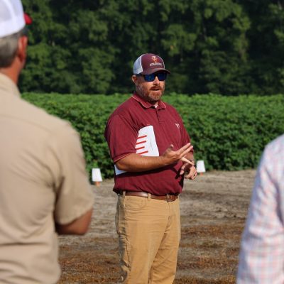 A man in baseball cap and polo shirt speaks to attendees at the cotton field day.