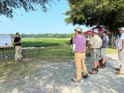 Man in sunglasses and black polo shirt speaks to a group of field day attendees outside.