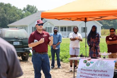 A man in hat and sunglasses addresses the crowd at cotton field day as students look on from behind him.