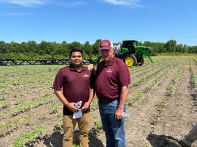 Two men in maroon shirts stand in the field with John Deere's See and Spray equipment in the background.