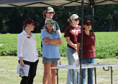 A group of field day attendees gathered beneath the shade of a canopy near a field of peanuts.