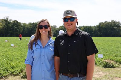 A woman and man wearing sunglasses smile while standing beside a field of peanuts.