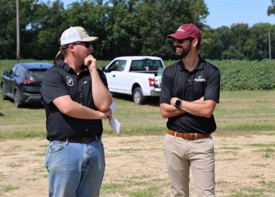 Two men in hats and sunglasses discussing field day events in field with trucks in background.