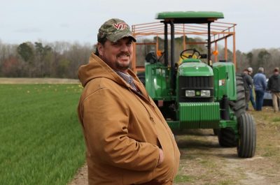 Man in jacket and baseball cap stands in front of tractor in the field during the Cover Crop Field Tour at Tidewater AREC.