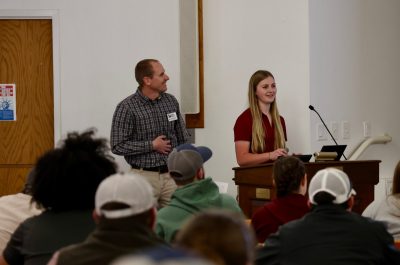 A young woman with long, blonde hair speaks to attendees from behind a podium during cover crop field day