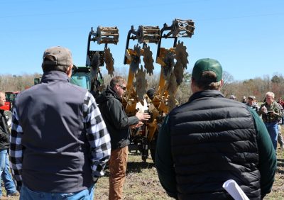 A man in a black jacket stands beside soil tillage equipment and he talks to attendees in the field during cover crop field day