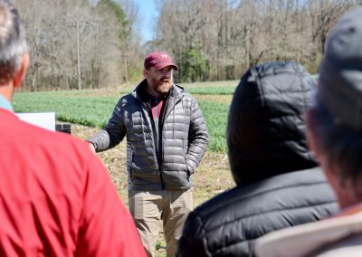 A man wearing a gray jacket and burgundy baseball cap speaks to attendees in the field during the cover crop field day event.