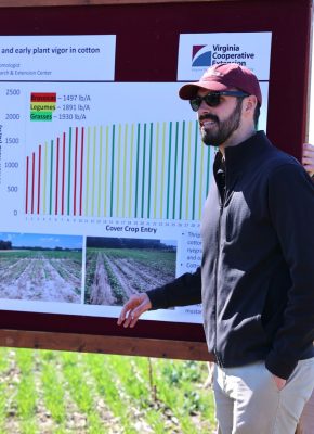 A man wearing dark sunglasses and a burgundy baseball cap stands in front of a bulletin board of data posters.