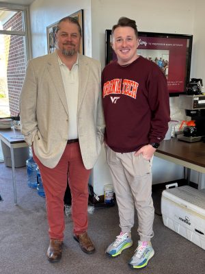 A man in a beige blazer stands beside a PhD student wearing a Virginia Tech sweatshirt in the lobby of the Tidewater AREC