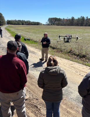 A man speaks to a group of field day attendees at the edge of a field with a large drone behind him prepared for flight.
