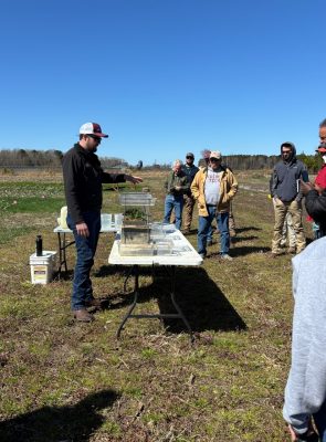 A man wearing a hat speaks to a group in a field as he stands behind a table with cover crop displays