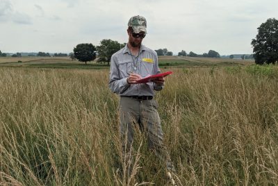 Assistant Professor Leighton Reid stands in the middle of a tall, grassy field, writing on a clipboard.